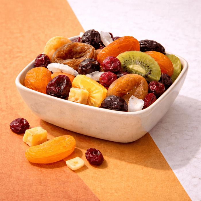 Bowl of premium mixed dried fruit including apricots, cranberries, dates, kiwi, banana chips and citrus pieces on a neutral background.